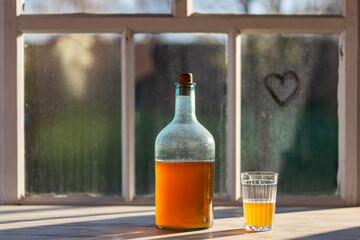 Big bottle with a drink made from fermented birch sap on the windowsill on a warm spring day, closeup. Traditional Ukrainian cold barley drink kvass in a glass jar and glass on table near yard - 782914454