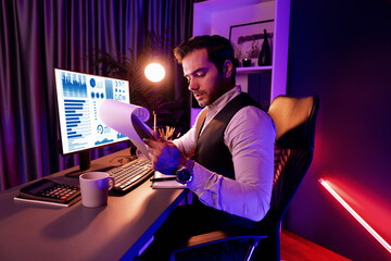 Serious smart businessman focusing on paperwork report folder analyzing sales channel discipline's product service comparing graph on pc screen market target customer at neon dark light room. Surmise. - obrazy, fototapety, plakaty