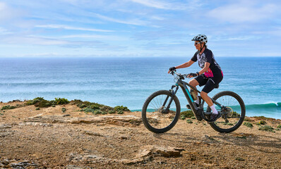 happy active senior woman cycling at the the rock cliffs and lighthouse of Cabo Sao Vicente, the south-western spit of Europe at the atlantic coast of  Algarve, Portugal, 