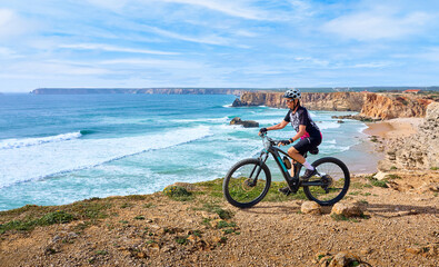 happy active senior woman cycling at the the rock cliffs and lighthouse of Cabo Sao Vicente, the south-western spit of Europe at the atlantic coast of  Algarve, Portugal,  - 782924624