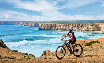 happy active senior woman cycling at the the rock cliffs and lighthouse of Cabo Sao Vicente, the south-western spit of Europe at the atlantic coast of  Algarve, Portugal,  - 782924873