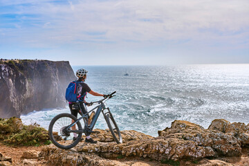 happy active senior woman cycling at the the rock cliffs and lighthouse of Cabo Sao Vicente, the south-western spit of Europe at the atlantic coast of  Algarve, Portugal,  - 782925090