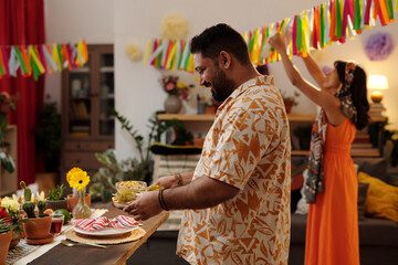 Side view of happy Hispanic man in Mexican shirt bringing homemade snacks for festive table against his girlfriend decorating living room - obrazy, fototapety, plakaty