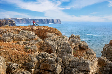 happy active senior woman cycling at the the rock cliffs and lighthouse of Cabo Sao Vicente, the south-western spit of Europe at the atlantic coast of Algarve, Portugal, 