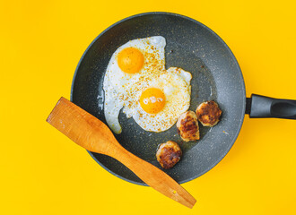 Two fried eggs in a frying pan next to three cutlets on a yellow background. Bachelor breakfast, lunch and dinner concept.