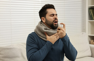 Young man with scarf using throat spray indoors - obrazy, fototapety, plakaty