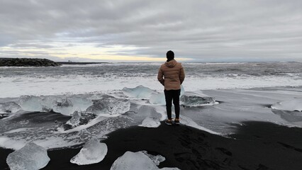 Man admiring the view of sea with foamy waves crashing on a black sandy beach with small icebergs