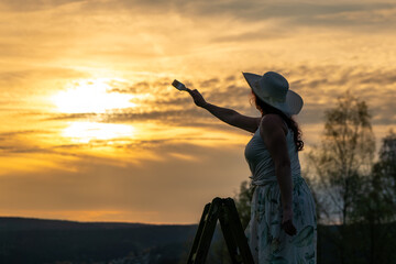a woman in a dress stands on a wooden ladder and paints the sunset
