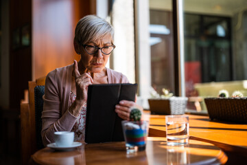 Mature woman is sitting in cafe and using digital tablet. - 782942271