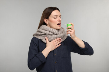 Woman with scarf using throat spray on grey background