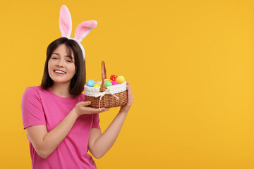 Easter celebration. Happy woman with bunny ears and wicker basket full of painted eggs on orange...