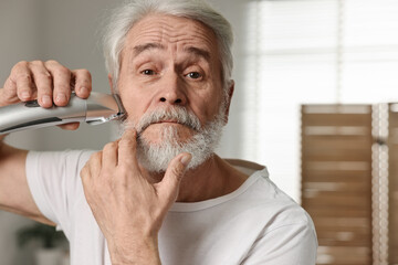 Senior man trimming beard with electric trimmer in bathroom