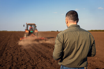 Rear view of mature farmer standing in field looking at tractor while cultivate the land.