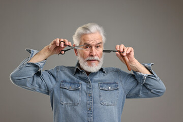 Senior man with mustache holding blade and scissors on grey background