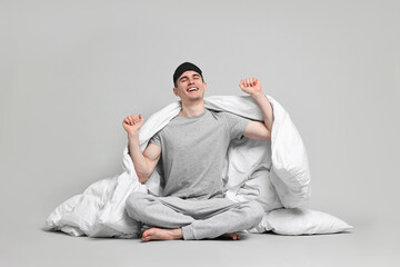 Happy man in pyjama and sleep mask wrapped in blanket on grey background