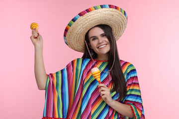 Young woman in Mexican sombrero hat and poncho dancing with maracas on pink background