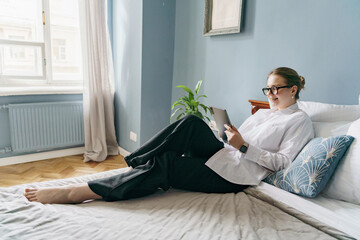 A relaxed professional woman reclines on a bed, engaging with a tablet, merging leisure with...