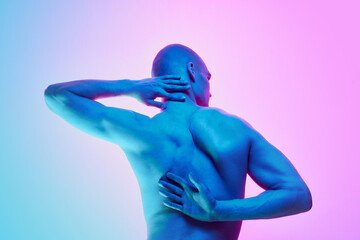 Back view of shirtless young bald man standing shirtless, showing muscular relief back body on gradient blue pink background in neon light. Concept of male beauty, body, youth, fitness, sport, health
