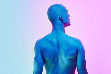Back view of shirtless young bald man standing shirtless, showing muscular relief back body on gradient blue pink background in neon light. Concept of male beauty, body, youth, fitness, sport, health
