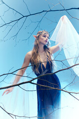 A young woman in a flowing blue dress, resembling an elf princess, stands gracefully atop a tree in a whimsical and fairy-like manner.