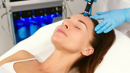 Fototapeta na wymiar A middle-aged woman receives facial treatment in a professional beauty salon. A close-up cosmetologist manipulates a hydropeeling device to clean and rejuvenate facial skin.