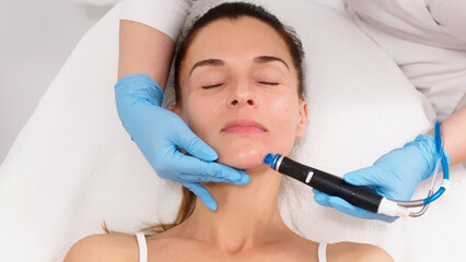 Naklejka premium A middle-aged woman receives facial treatment in a professional beauty salon. A close-up cosmetologist manipulates a hydropeeling device to clean and rejuvenate facial skin.