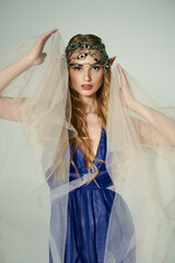 A young woman exudes elegance in a blue dress with a delicate veil over her head, embodying the essence of a mystical fairy princess.