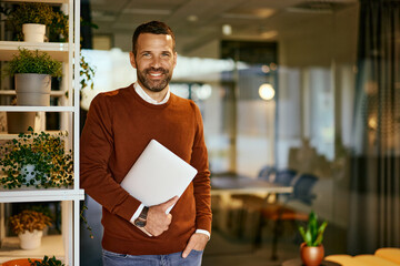 Portrait of a smiling businessman, holding a laptop, posing for the camera.