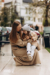 little happy girl dressed in a coat sits on her mother's arms against the background of the city