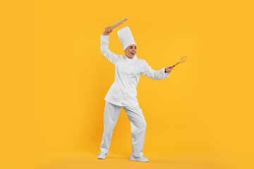 Happy professional confectioner in uniform holding rolling pin and whisk on yellow background