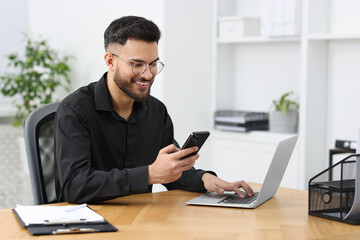 Handsome young man using smartphone while working with laptop at wooden table in office