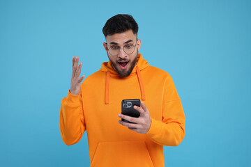 Shocked young man using smartphone on light blue background