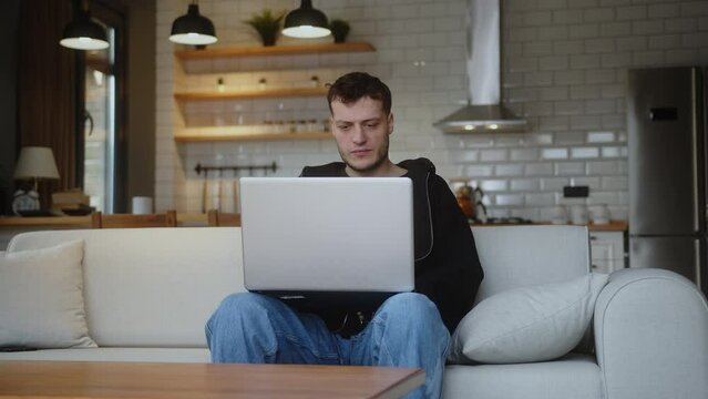 Young man sit on sofa using laptop watching video, surfing internet, using social media, chatting with friends. Leisure activity, spend free time on internet in living room at home