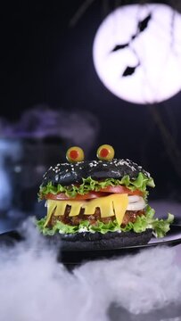 Monster burger. Black bun, beef cutlet, lettuce, onion, tomato and cheese in the shape of teeth, Against the backdrop of the moon and a steaming cauldron. Slow, vertical motion, circular rotation