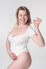 Happiness plus size woman in white bodysuit shows OK sign. Extended sizes mature adult model smiling and looking at camera posing on white background. Generation X curvy fashion model. Body positive.