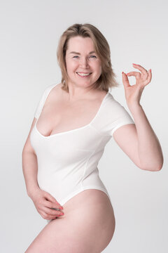 Happiness plus size woman in white bodysuit shows OK sign. Extended sizes mature adult model smiling and looking at camera posing on white background. Generation X curvy fashion model. Body positive.