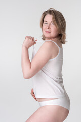 Middle aged overweight woman wearing white underwear, holding strap of camisole and placing hand on her stomach. Caucasian Generation X woman represents the concept of a healthy lifestyle for ladies
