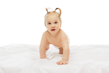 Little cute baby girl, child in diaper, with two ponytails crawling on white bedsheets with curious expression against white background. Concept of childhood, care, health, well-being, parenthood