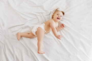 Active, smiling, little redhead girl, toddler in diaper lying on belly on bed, laughing, playing on white bedsheets. Happiness, fun and joy. Concept of childhood, care, health, well-being, parenthood