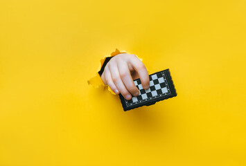 A right child's hand emerges from a torn hole in yellow paper and holds a small chessboard. The...
