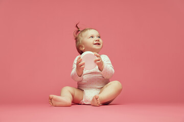 Smiling baby in comfortable onesie sitting on pink background, holding moisturizing skin cream. Cosmetics for babies. Concept of childhood, care, health, well-being, parenthood