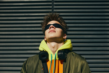 Portrait of man in trendy streetwear, neon hoodie, sunglasses, headphones posing against metal slatted background. Concept of street fashion and urban style, modern lifestyle, gen Z, self-expression. - obrazy, fototapety, plakaty