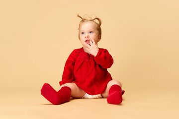 Little baby girl, child with ponytails in red dress and socks sitting against beige studio background. Concept of childhood, care, health, well-being, parenthood