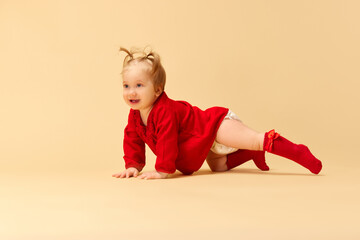 Little baby girl, child with ponytails in red dress and socks crawling, playing against beige studio background. Concept of childhood, care, health, well-being, parenthood