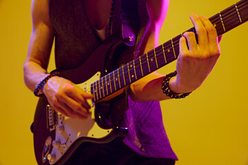 Close up photo of young bass guitarist's hands playing drive, energetic music in neon light against yellow studio background. Concept of music and art, hobby, concerts and festivals, modern culture