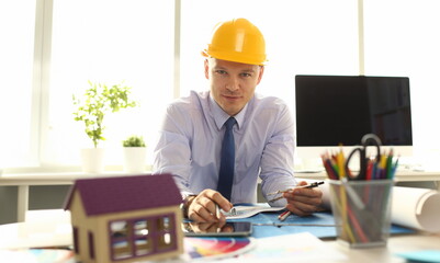 Confident Architect Designer Create Building Plan. Handsome Man in Shirt and Yellow Helmet Designing House Model on Blueprint. Safety Constracting Concept. Stationery, Instruments on Worktop