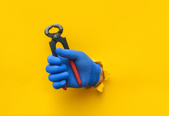A right man's hand in a blue fabric knitted glove holds wire cutters. Torn hole in yellow paper....