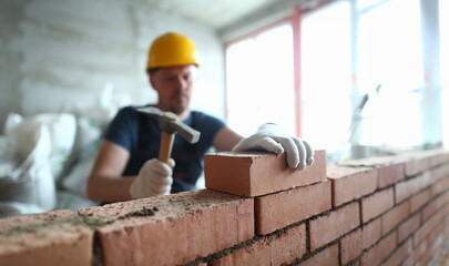 Portrait of skilled man constructing big concrete wall and using special hammer to properly fulfill task to gently lay bricks on unfinished structure. Building concept - obrazy, fototapety, plakaty