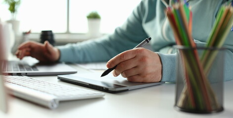 A young designer holds pen from tablet in hand working on new project for customer in field of...