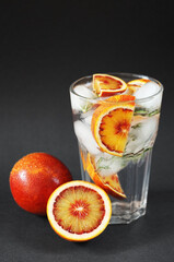Glass with water, ice and citrus next to blood oranges on a dark background